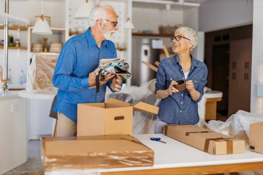 Un couple âgé se tient dans une cuisine moderne, souriant alors qu'ils préparent leur tri affaires déménagement retraité. La femme tient une tasse, l'homme emballe des objets dans du papier journal et les affaires déballées reposent sur le comptoir.
