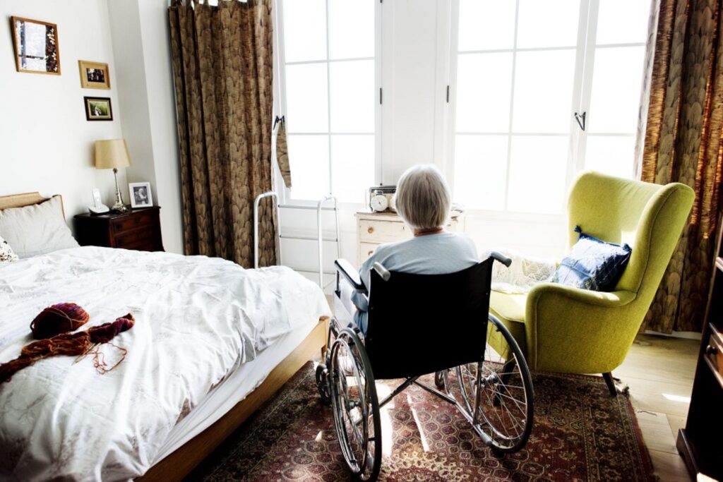 Une personne âgée aux cheveux gris courts est assise dans un fauteuil roulant, face aux grandes fenêtres d'une chambre douillette baignée de lumière naturelle. Elle réfléchit tranquillement au récent refus d'aide senior et à ses sentiments concernant l'aide à domicile imposée.