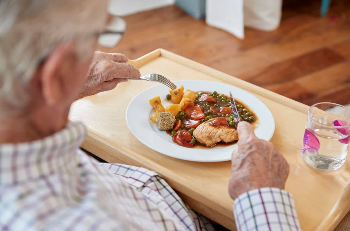 Une personne âgée est assise à une table, dégustant un repas composé de poulet, de pommes de terre, de petits pois et de carottes dans une assiette blanche - peut-être dans le cadre du repas domicile PACA - avec un verre d'eau à proximité, à l'aide d'un couteau et d'une fourchette.