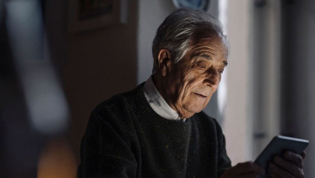 Un homme âgé aux cheveux gris, vêtu d'un pull sombre, est assis à l'intérieur ; il tient une tablette qu'il regarde d'un air pensif, peut-être en train de lire des informations sur l'ostéoporose chez les personnes âgées ou sur les moyens de prévenir les fractures du col du fémur chez les seniors, tandis qu'une douce lumière illumine son visage.