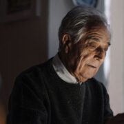 Un homme âgé aux cheveux gris, vêtu d'un pull sombre, est assis à l'intérieur ; il tient une tablette qu'il regarde d'un air pensif, peut-être en train de lire des informations sur l'ostéoporose chez les personnes âgées ou sur les moyens de prévenir les fractures du col du fémur chez les seniors, tandis qu'une douce lumière illumine son visage.