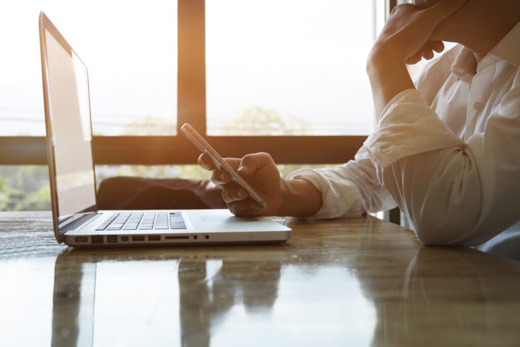 Une personne en chemise blanche est assise à un bureau avec un ordinateur portable ouvert, tenant un smartphone - peut-être en train de coordonner l'aide à domicile à distance - reposant son menton sur sa main tandis que la lumière du soleil traverse de grandes fenêtres à l'arrière-plan.
