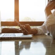 Une personne en chemise blanche est assise à un bureau avec un ordinateur portable ouvert, tenant un smartphone - peut-être en train de coordonner l'aide à domicile à distance - reposant son menton sur sa main tandis que la lumière du soleil traverse de grandes fenêtres à l'arrière-plan.
