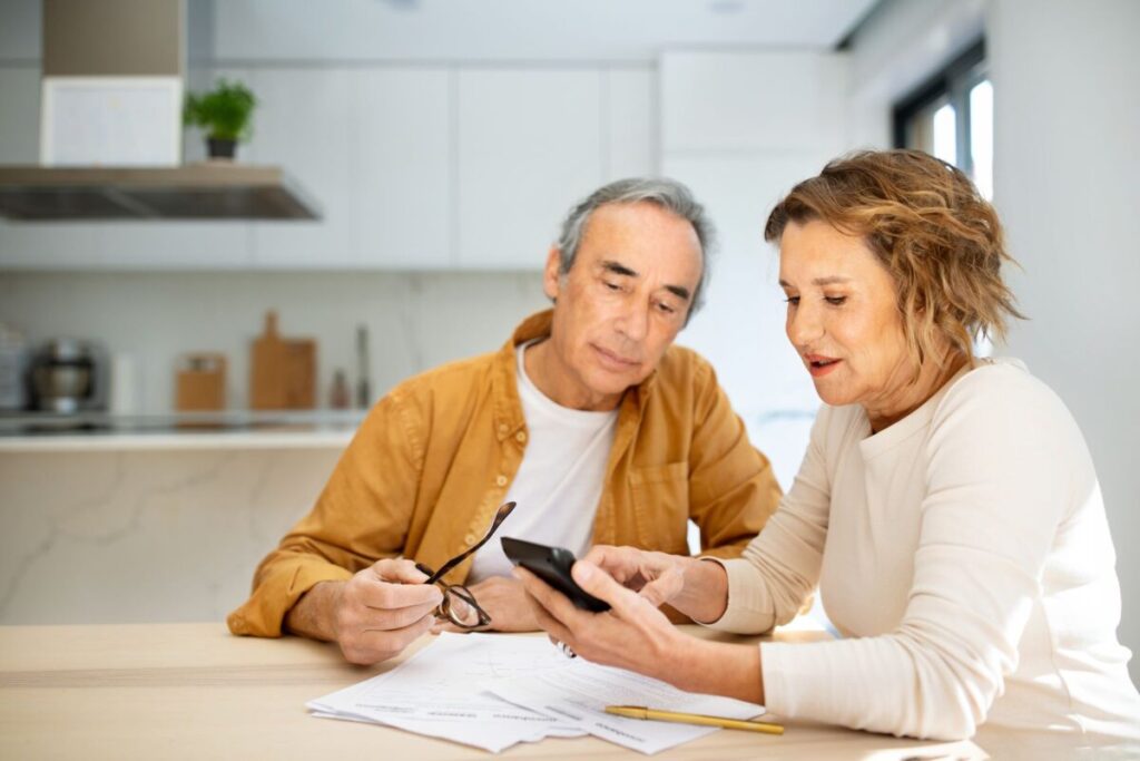 Un couple âgé est assis à la table de la cuisine, en train d'examiner les documents relatifs à la demande de CMI pour personnes âgées. La femme tient un smartphone et montre l'écran du doigt, tandis que l'homme l'observe, ses lunettes à la main. Tous deux semblent concentrés et attentifs.