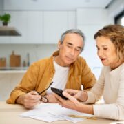Un couple âgé est assis à la table de la cuisine, en train d'examiner les documents relatifs à la demande de CMI pour personnes âgées. La femme tient un smartphone et montre l'écran du doigt, tandis que l'homme l'observe, ses lunettes à la main. Tous deux semblent concentrés et attentifs.