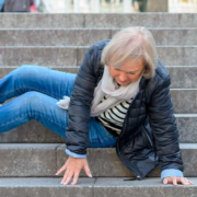 Une femme âgée, vêtue d'une veste noire et d'un jean bleu, est assise sur les marches d'un escalier extérieur ; on dirait qu'elle est tombée. Elle s'appuie sur ses mains et regarde vers le bas, l'air surpris, ce qui met en évidence le besoin de mesures visant à renforcer le lien social auprès des personnes âgées.