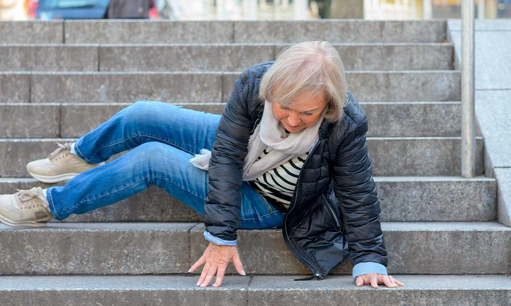 Une femme âgée, vêtue d'une veste noire et d'un jean bleu, est assise sur les marches d'un escalier extérieur ; on dirait qu'elle est tombée. Elle s'appuie sur ses mains et regarde vers le bas, l'air surpris, ce qui met en évidence le besoin de mesures visant à renforcer le lien social auprès des personnes âgées.