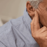 Gros plan d'un homme âgé aux cheveux grisonnants, se touchant l'oreille et regardant vers le bas en signe d'inconfort - peut-être atteint de presbyacousie senior, qui peut être détectée précocement grâce au dépistage audition senior. Il porte une chemise bleu clair.