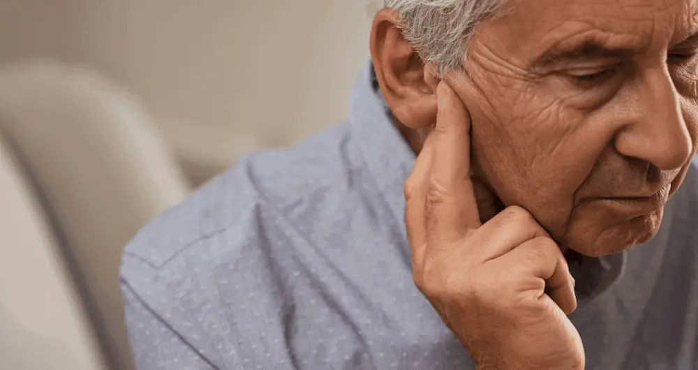 Gros plan d'un homme âgé aux cheveux grisonnants, se touchant l'oreille et regardant vers le bas en signe d'inconfort - peut-être atteint de presbyacousie senior, qui peut être détectée précocement grâce au dépistage audition senior. Il porte une chemise bleu clair.