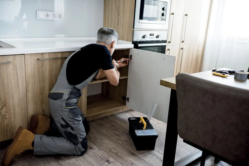 Un homme en salopette est agenouillé par terre, en train de réparer la porte d'un placard dans une cuisine moderne — l'idéal pour ceux qui souhaitent monter des meubles chez eux et profiter du crédit d'impôt bricolage jusqu'au plafond de 500 € avec un homme à tout faire.