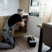 Un homme en salopette est agenouillé par terre, en train de réparer la porte d'un placard dans une cuisine moderne — l'idéal pour ceux qui souhaitent monter des meubles chez eux et profiter du crédit d'impôt bricolage jusqu'au plafond de 500 € avec un homme à tout faire.