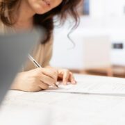 Une femme est assise à une table, en train de signer au stylo un document relatif à la grille AGGIR, tandis qu’une autre personne se tient debout à proximité. Des papiers et un ordinateur portable sont visibles sur la table, soulignant l’importance accordée à cette étape clé de l’accompagnement médico-social.