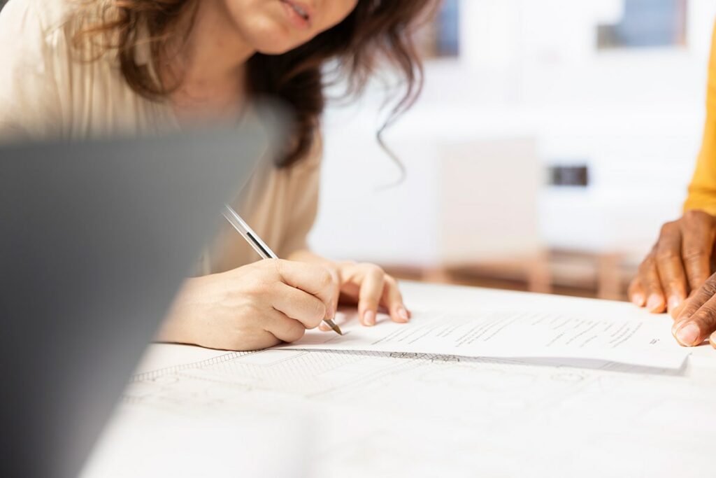Une femme est assise à une table, en train de signer au stylo un document relatif à la grille AGGIR, tandis qu’une autre personne se tient debout à proximité. Des papiers et un ordinateur portable sont visibles sur la table, soulignant l’importance accordée à cette étape clé de l’accompagnement médico-social.