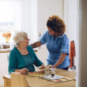 Une aide-soignante en uniforme bleu sert le petit-déjeuner à une femme âgée souriante, assise à la table de la cuisine ; elle l'aide à suivre son traitement d'oxygénothérapie à domicile et crée une ambiance chaleureuse et conviviale.