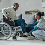 Une infirmière en blouse s'agenouille pour aider un homme souriant en fauteuil roulant à lacer ses chaussures dans un salon lumineux — un exemple de « projet d'accompagnement personnalisé » dans le cadre de l'aide à domicile, où les soins sont adaptés aux besoins de chacun.