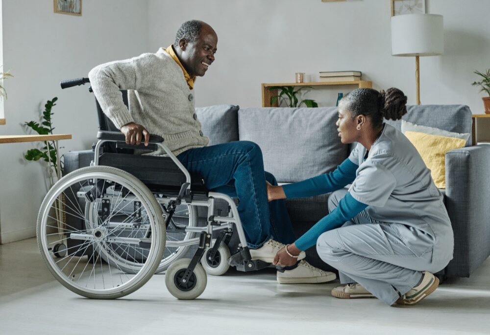 Une infirmière en blouse s'agenouille pour aider un homme souriant en fauteuil roulant à lacer ses chaussures dans un salon lumineux — un exemple de « projet d'accompagnement personnalisé » dans le cadre de l'aide à domicile, où les soins sont adaptés aux besoins de chacun.