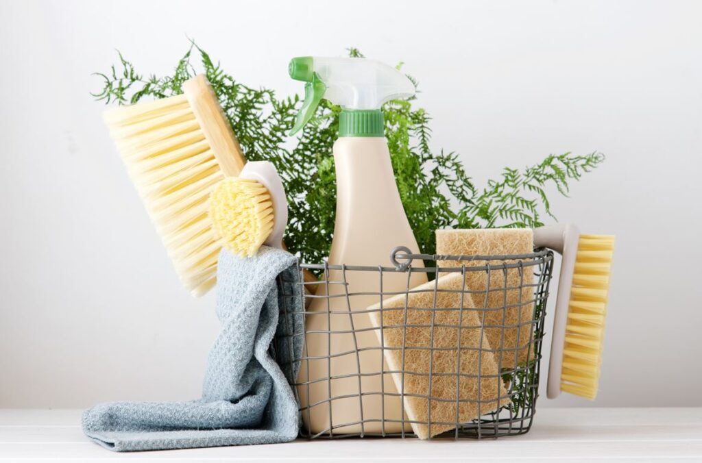 Un panier en fil de fer rempli de produits ménagers naturels — tels que des brosses, un vaporisateur, des éponges et un chiffon bleu — est posé sur une surface blanche, avec des plantes vertes en arrière-plan. Idéal pour une recette de nettoyant multi-usage fait maison.
