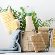 Un panier en fil de fer rempli de produits ménagers naturels — tels que des brosses, un vaporisateur, des éponges et un chiffon bleu — est posé sur une surface blanche, avec des plantes vertes en arrière-plan. Idéal pour une recette de nettoyant multi-usage fait maison.