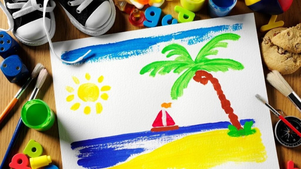 Une peinture d'enfant aux couleurs vives représente une scène de plage avec un palmier, le soleil, la mer bleue et un voilier rouge. Entourée de matériel de dessin, de biscuits et de jouets posés sur une table en bois : une façon idéale d'occuper un enfant pendant les vacances d'été.