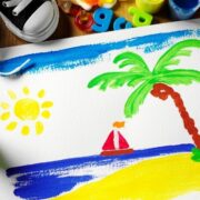 Une peinture d'enfant aux couleurs vives représente une scène de plage avec un palmier, le soleil, la mer bleue et un voilier rouge. Entourée de matériel de dessin, de biscuits et de jouets posés sur une table en bois : une façon idéale d'occuper un enfant pendant les vacances d'été.