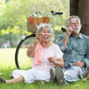 Un couple de personnes âgées est assis sur l'herbe, à l'ombre d'un arbre ; ils font des bulles de savon et sourient, incarnant ainsi « le bien vieillir après 60 ans ». Derrière eux se trouve un vélo muni d'un panier. C'est une journée ensoleillée dans un parc, avec des arbres verdoyants en arrière-plan, ce qui met en évidence l'importance du lien social pour les personnes âgées.