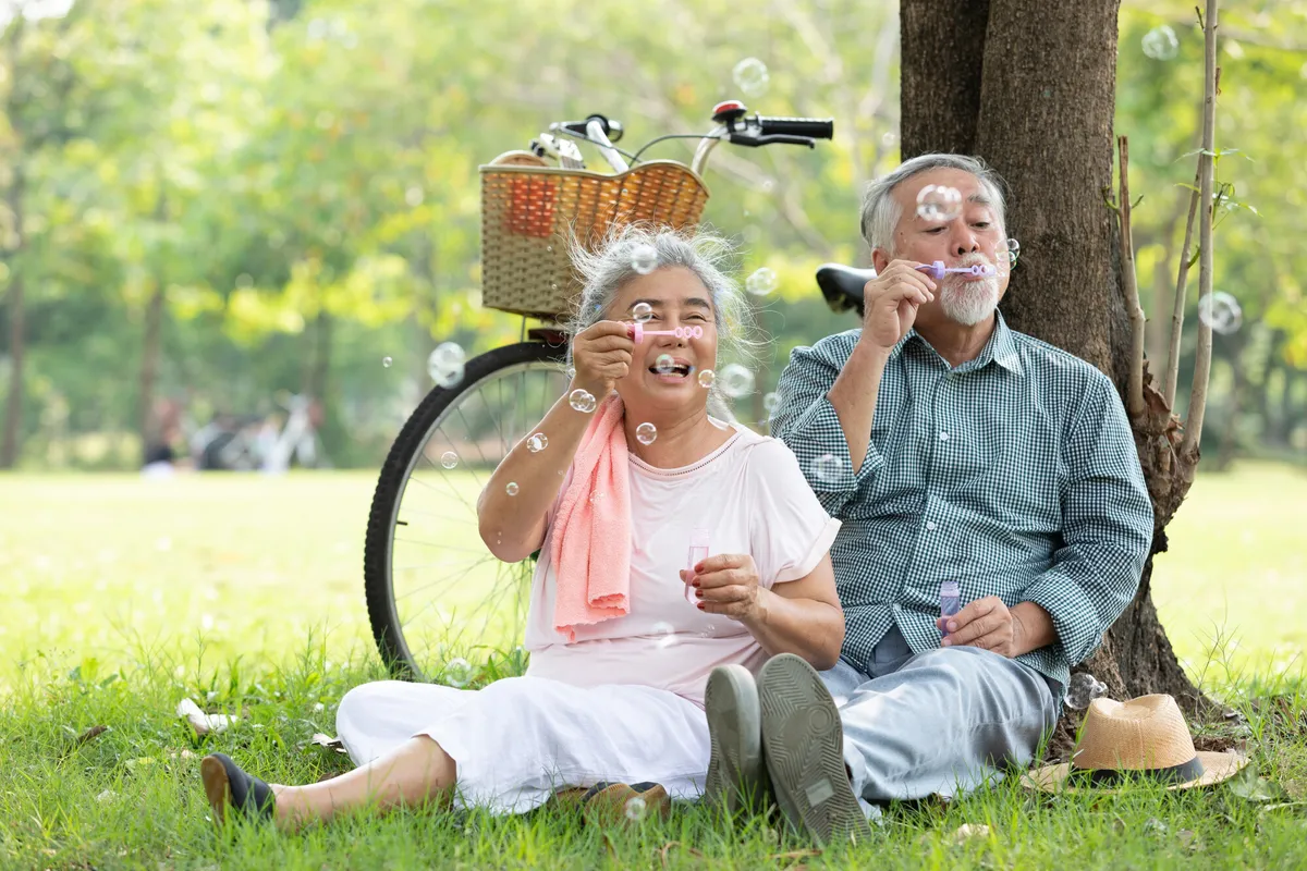 Un couple de personnes âgées est assis sur l'herbe, à l'ombre d'un arbre ; ils font des bulles de savon et sourient, incarnant ainsi « le bien vieillir après 60 ans ». Derrière eux se trouve un vélo muni d'un panier. C'est une journée ensoleillée dans un parc, avec des arbres verdoyants en arrière-plan, ce qui met en évidence l'importance du lien social pour les personnes âgées.