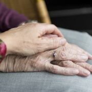 La main d’une personne âgée, ornée d’une bague, repose sur ses genoux tandis qu’une autre main s’y pose délicatement, lui apportant réconfort et soutien — un moment intime que l’on observe souvent lors de la recherche de signes de fragilité chez les personnes âgées ou lors d’une visite de contrôle chez un parent âgé visant à s’assurer de son bien-être.