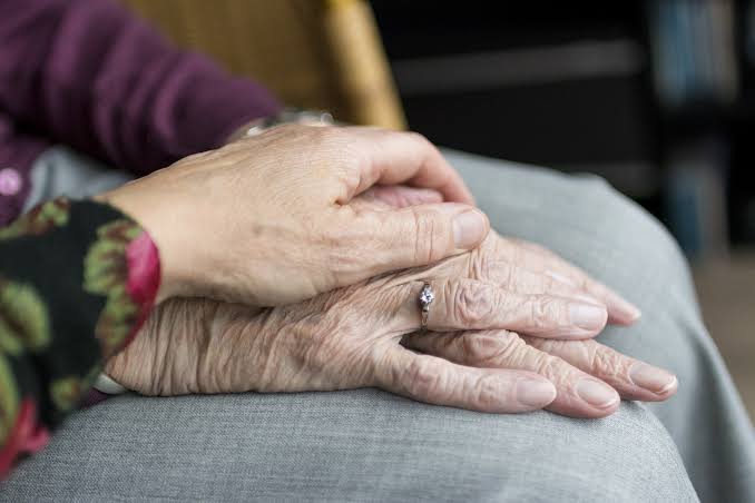 La main d’une personne âgée, ornée d’une bague, repose sur ses genoux tandis qu’une autre main s’y pose délicatement, lui apportant réconfort et soutien — un moment intime que l’on observe souvent lors de la recherche de signes de fragilité chez les personnes âgées ou lors d’une visite de contrôle chez un parent âgé visant à s’assurer de son bien-être.