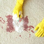 Deux mains gantées de jaune nettoient une tache de liquide rouge sur un tapis clair à l'aide d'une éponge et d'une mousse nettoyante, illustrant ainsi comment le maintien de la propreté peut constituer une alternative efficace à l'EHPAD pour les personnes âgées qui préfèrent rester chez elles.
