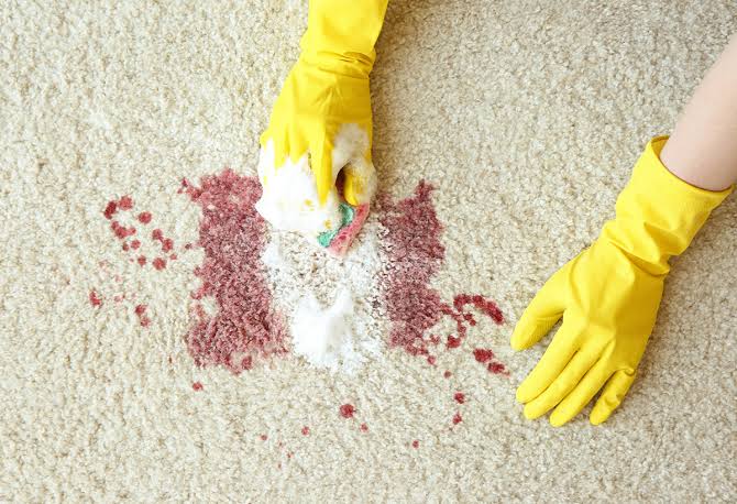 Deux mains gantées de jaune nettoient une tache de liquide rouge sur un tapis clair à l'aide d'une éponge et d'une mousse nettoyante, illustrant ainsi comment le maintien de la propreté peut constituer une alternative efficace à l'EHPAD pour les personnes âgées qui préfèrent rester chez elles.