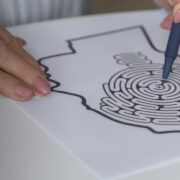 Une personne qui résout, à l'aide d'un stylo, un labyrinthe en forme de tête humaine, en se concentrant sur le centre du labyrinthe : une façon captivante de stimuler la mémoire des seniors. La feuille de papier repose sur une table blanche.
