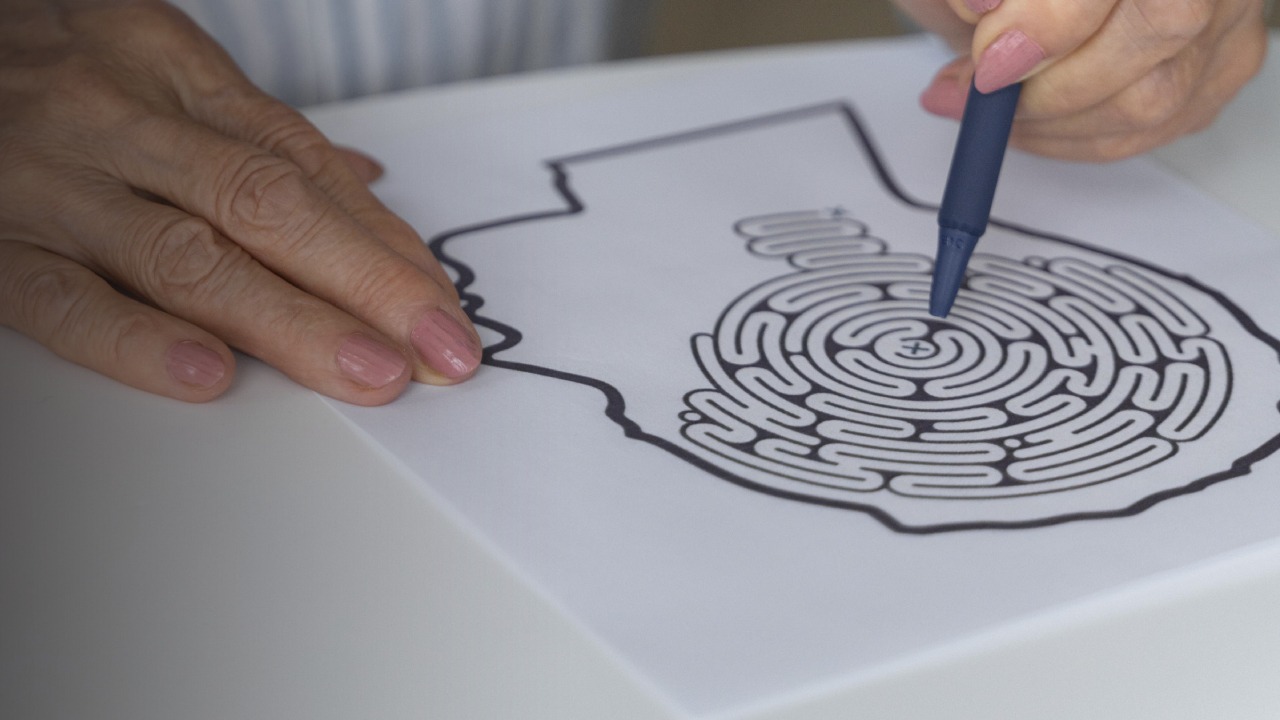 Une personne qui résout, à l'aide d'un stylo, un labyrinthe en forme de tête humaine, en se concentrant sur le centre du labyrinthe : une façon captivante de stimuler la mémoire des seniors. La feuille de papier repose sur une table blanche.
