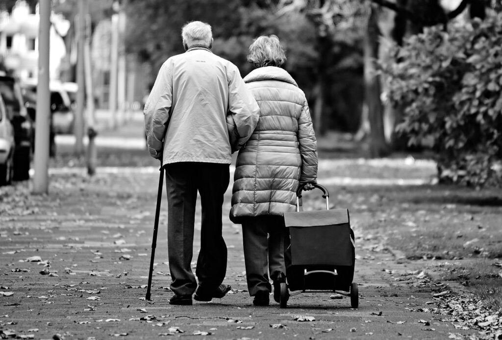 Un couple de personnes âgées marche le long d'un sentier bordé d'arbres. L'homme s'appuie sur une canne et la femme tire un caddie. Des feuilles mortes recouvrent le sol. Cette image en noir et blanc évoque la résilience discrète des personnes âgées qui pourraient bénéficier d'une inscription au registre communal des personnes vulnérables, une mesure importante pour mieux protéger chaque personne âgée isolée.