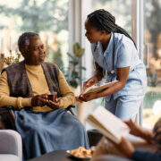 Une infirmière sourit et discute avec un homme âgé en fauteuil roulant qui tient un verre, tandis qu'une autre femme est assise à proximité et lit un livre. La scène se déroule à l'intérieur, dans une pièce lumineuse dotée de grandes fenêtres, illustrant la chaleur et le confort que peuvent offrir ces structures, en alternative à l'EHPAD, aux personnes âgées à la recherche de soins personnalisés à domicile.