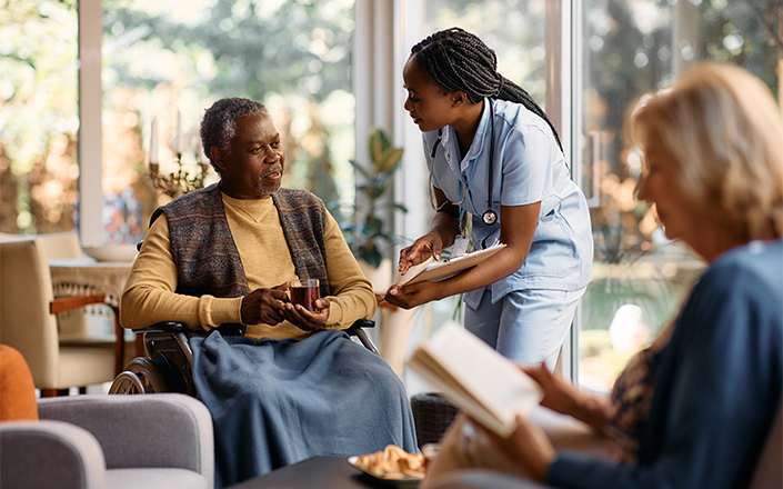 Une infirmière sourit et discute avec un homme âgé en fauteuil roulant qui tient un verre, tandis qu'une autre femme est assise à proximité et lit un livre. La scène se déroule à l'intérieur, dans une pièce lumineuse dotée de grandes fenêtres, illustrant la chaleur et le confort que peuvent offrir ces structures, en alternative à l'EHPAD, aux personnes âgées à la recherche de soins personnalisés à domicile.