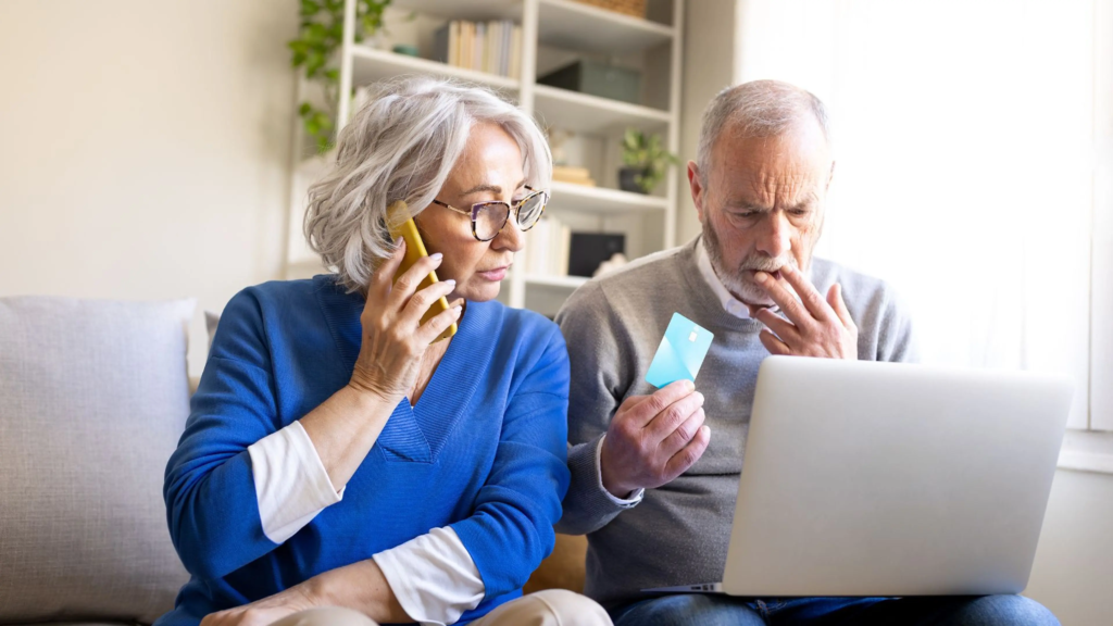 Un couple de personnes âgées est assis sur un canapé ; la femme est au téléphone, probablement avec un faux conseiller bancaire spécialisé dans les seniors, tandis que l'homme tient une carte de crédit et regarde d'un air inquiet l'écran d'un ordinateur portable, ce qui met en évidence la menace croissante que représentent les arnaques visant les personnes âgées.
