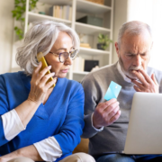Un couple de personnes âgées est assis sur un canapé ; la femme est au téléphone, probablement avec un faux conseiller bancaire spécialisé dans les seniors, tandis que l'homme tient une carte de crédit et regarde d'un air inquiet l'écran d'un ordinateur portable, ce qui met en évidence la menace croissante que représentent les arnaques visant les personnes âgées.