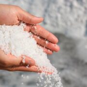 Une paire de mains tenant et versant du gros sel de mer blanc, avec davantage de sel et d'eau en arrière-plan flou — un rappel subtil que l'hyponatrémie peut survenir chez les personnes âgées en cas de consommation excessive d'eau, en particulier chez les seniors.