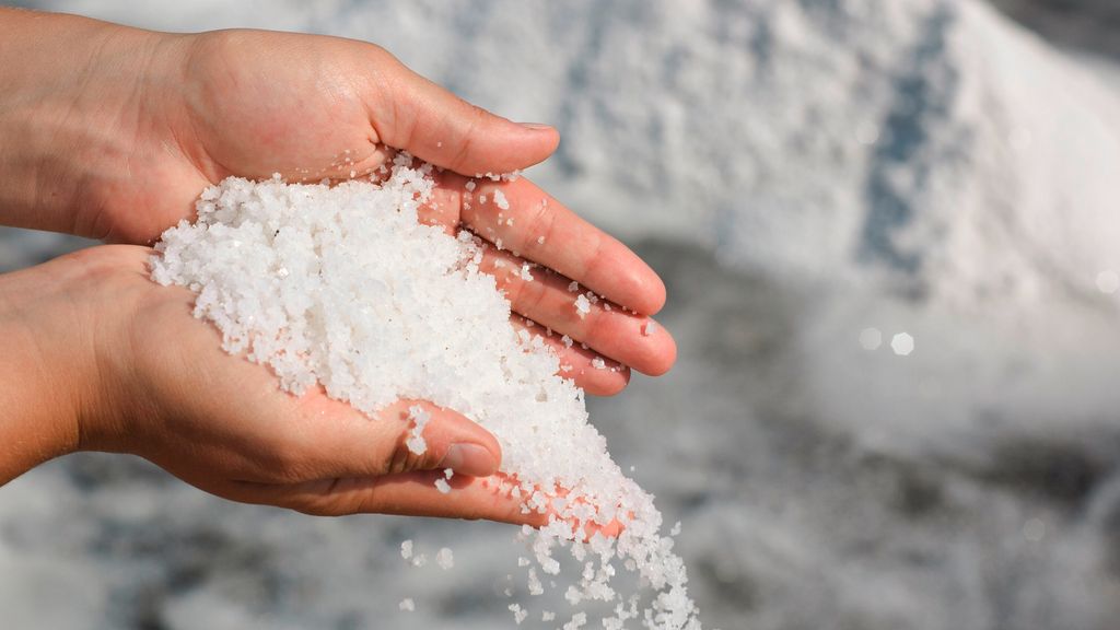 Une paire de mains tenant et versant du gros sel de mer blanc, avec davantage de sel et d'eau en arrière-plan flou — un rappel subtil que l'hyponatrémie peut survenir chez les personnes âgées en cas de consommation excessive d'eau, en particulier chez les seniors.