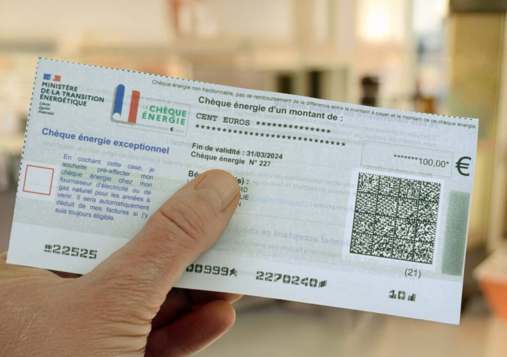 Une main tient un « Chèque Énergie » français d'une valeur de 100 euros, destiné à aider au paiement des factures d'énergie et soumis aux conditions d'utilisation du Chèque Énergie. Le chèque comporte les logos officiels, un code QR et les modalités d'utilisation. L'arrière-plan est flou, ce qui suggère un décor d'intérieur.