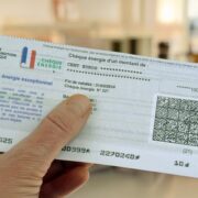 Une main tient un « Chèque Énergie » français d'une valeur de 100 euros, destiné à aider au paiement des factures d'énergie et soumis aux conditions d'utilisation du Chèque Énergie. Le chèque comporte les logos officiels, un code QR et les modalités d'utilisation. L'arrière-plan est flou, ce qui suggère un décor d'intérieur.