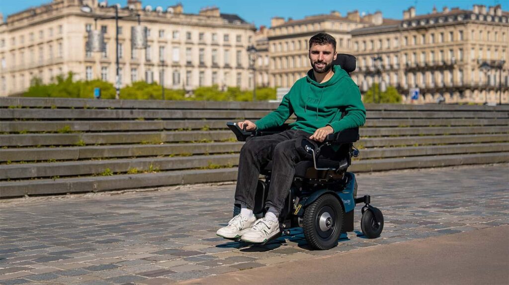 Un homme vêtu d'un sweat à capuche vert sourit alors qu'il se déplace en fauteuil roulant remis à neuf sur une allée pavée, avec en arrière-plan des bâtiments historiques et de la verdure.
