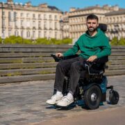 Un homme vêtu d'un sweat à capuche vert sourit alors qu'il se déplace en fauteuil roulant remis à neuf sur une allée pavée, avec en arrière-plan des bâtiments historiques et de la verdure.