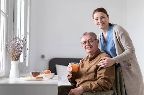 Un homme âgé est assis à une table, devant un jus de fruits et des fruits, et sourit. Une jeune femme, sans doute une aide-soignante, se tient derrière lui, les mains posées sur ses épaules, et sourit à la caméra. Leur bonheur reflète peut-être la sérénité que procure la participation à la campagne de vaccination contre la grippe et la Covid-19, qui garantit à tous deux une protection pendant cette saison.