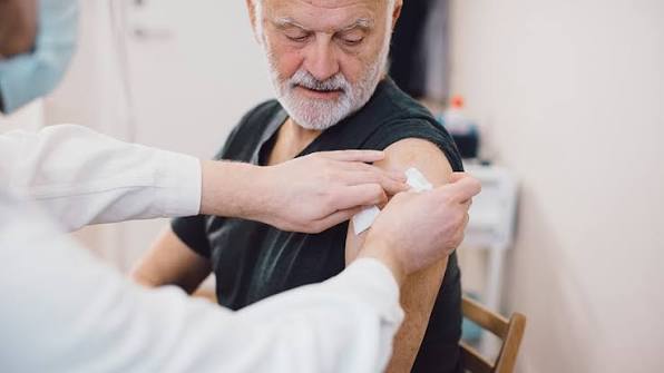 Un professionnel de santé essuie le haut du bras d'un homme âgé, assis et vêtu d'une chemise à manches courtes, afin de le préparer à une injection ou à une vaccination dans un établissement médical. Cette scène souligne l'importance de se faire vacciner lorsqu'on ne peut pas se déplacer, afin de garantir que même les personnes à mobilité réduite aient accès aux vaccinations essentielles, telles que la vaccination contre la grippe et la Covid.