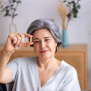 Une femme d'un certain âge aux cheveux gris, vêtue d'un t-shirt gris clair, tient à la main une gélule jaune et noire bien nette, soulignant ainsi l'importance de l'apport en vitamine D chez les personnes âgées. Des plantes d'ornement et un buffet en bois apparaissent en arrière-plan, flous.
