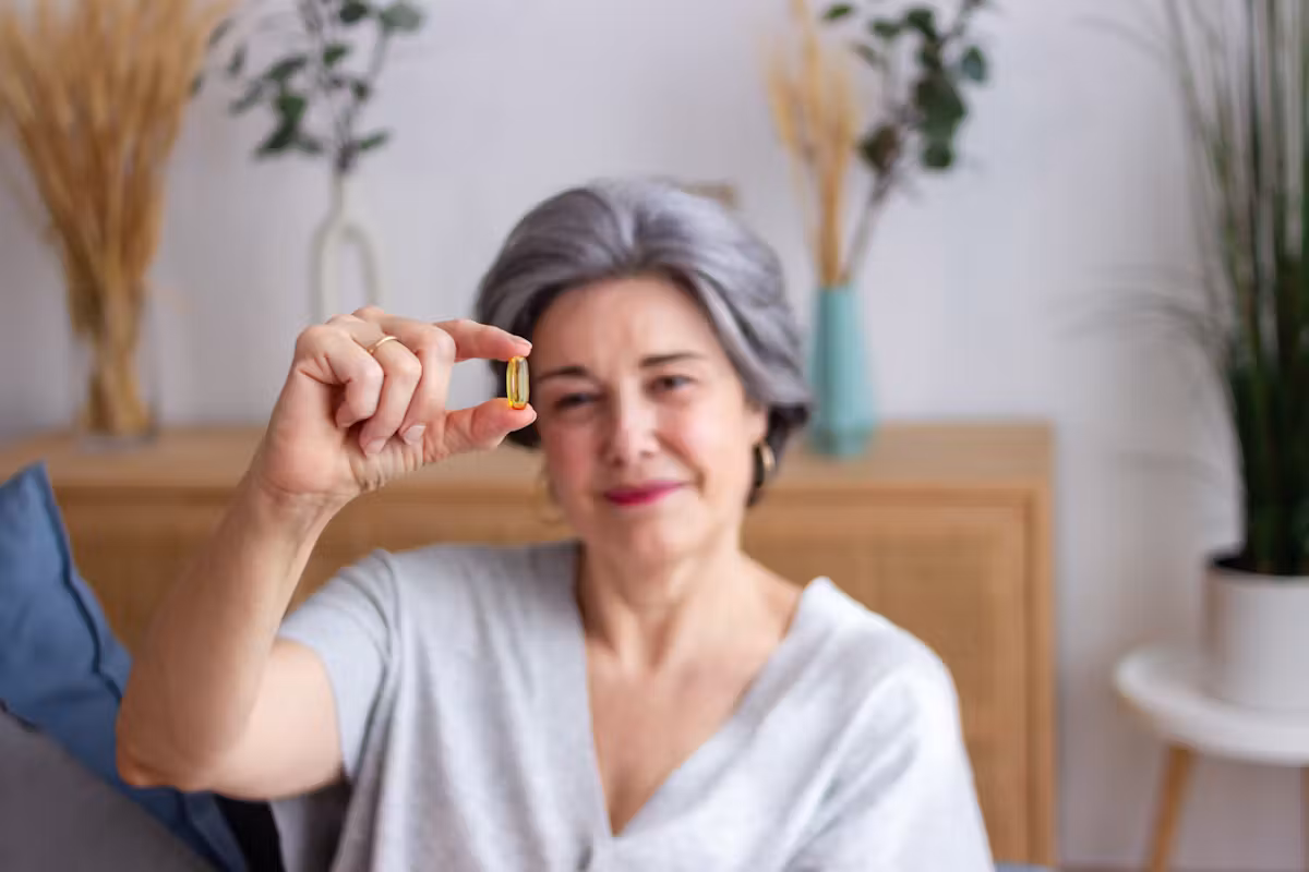 Une femme d'un certain âge aux cheveux gris, vêtue d'un t-shirt gris clair, tient à la main une gélule jaune et noire bien nette, soulignant ainsi l'importance de l'apport en vitamine D chez les personnes âgées. Des plantes d'ornement et un buffet en bois apparaissent en arrière-plan, flous.
