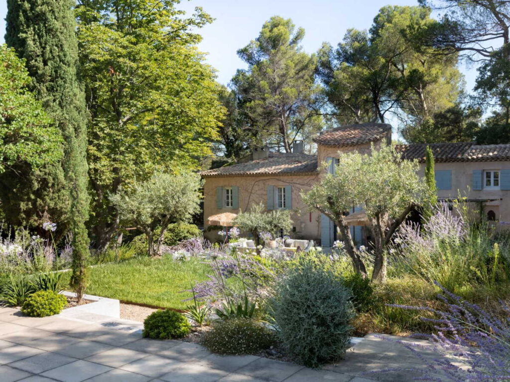 Une maison en pierre, entourée d’arbres luxuriants, d’arbustes fleuris et d’une pelouse verdoyante, baigne sous un ciel bleu limpide. Le jardin, agrémenté de lavande et de divers arbustes, offre un cadre paisible et ensoleillé — un environnement idéal pour réfléchir à la manière de financer l’aménagement de la salle de bains d’un parent ou pour se demander qui peut prescrire un fauteuil percé afin d’assurer le bien-être des résidents âgés.