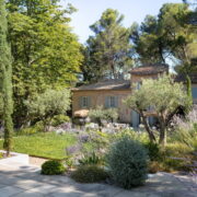 Une maison en pierre, entourée d’arbres luxuriants, d’arbustes fleuris et d’une pelouse verdoyante, baigne sous un ciel bleu limpide. Le jardin, agrémenté de lavande et de divers arbustes, offre un cadre paisible et ensoleillé — un environnement idéal pour réfléchir à la manière de financer l’aménagement de la salle de bains d’un parent ou pour se demander qui peut prescrire un fauteuil percé afin d’assurer le bien-être des résidents âgés.