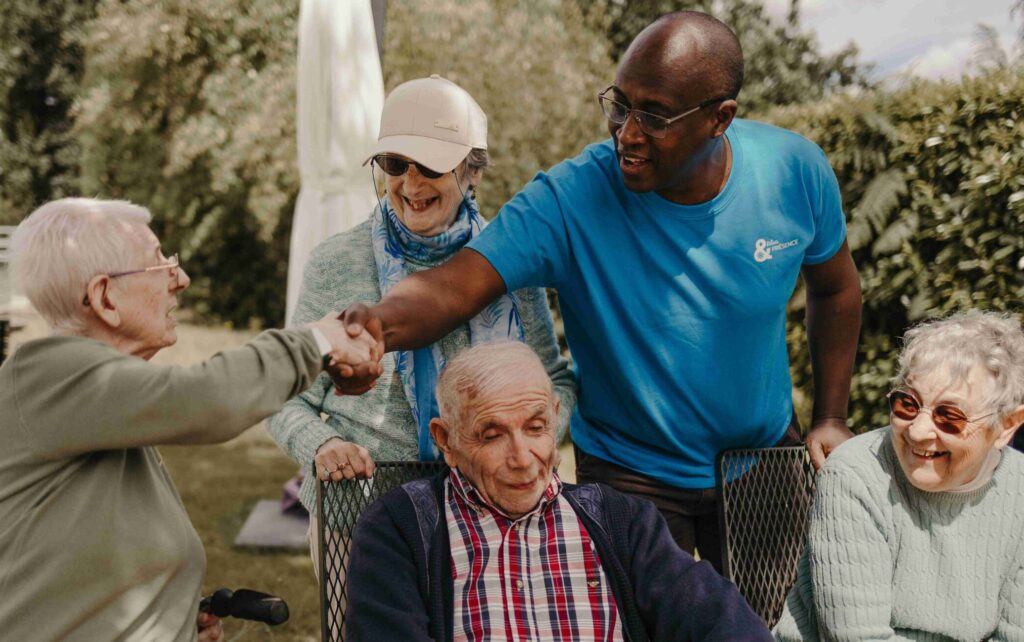 Un homme en chemise bleue serre la main d’un homme âgé à l’extérieur, tandis que trois autres personnes âgées sourient en les regardant. Ils sont réunis par une journée ensoleillée, au milieu de la verdure, illustrant ainsi l’esprit de solidarité qui est au cœur de la Journée nationale des aidants 2026.